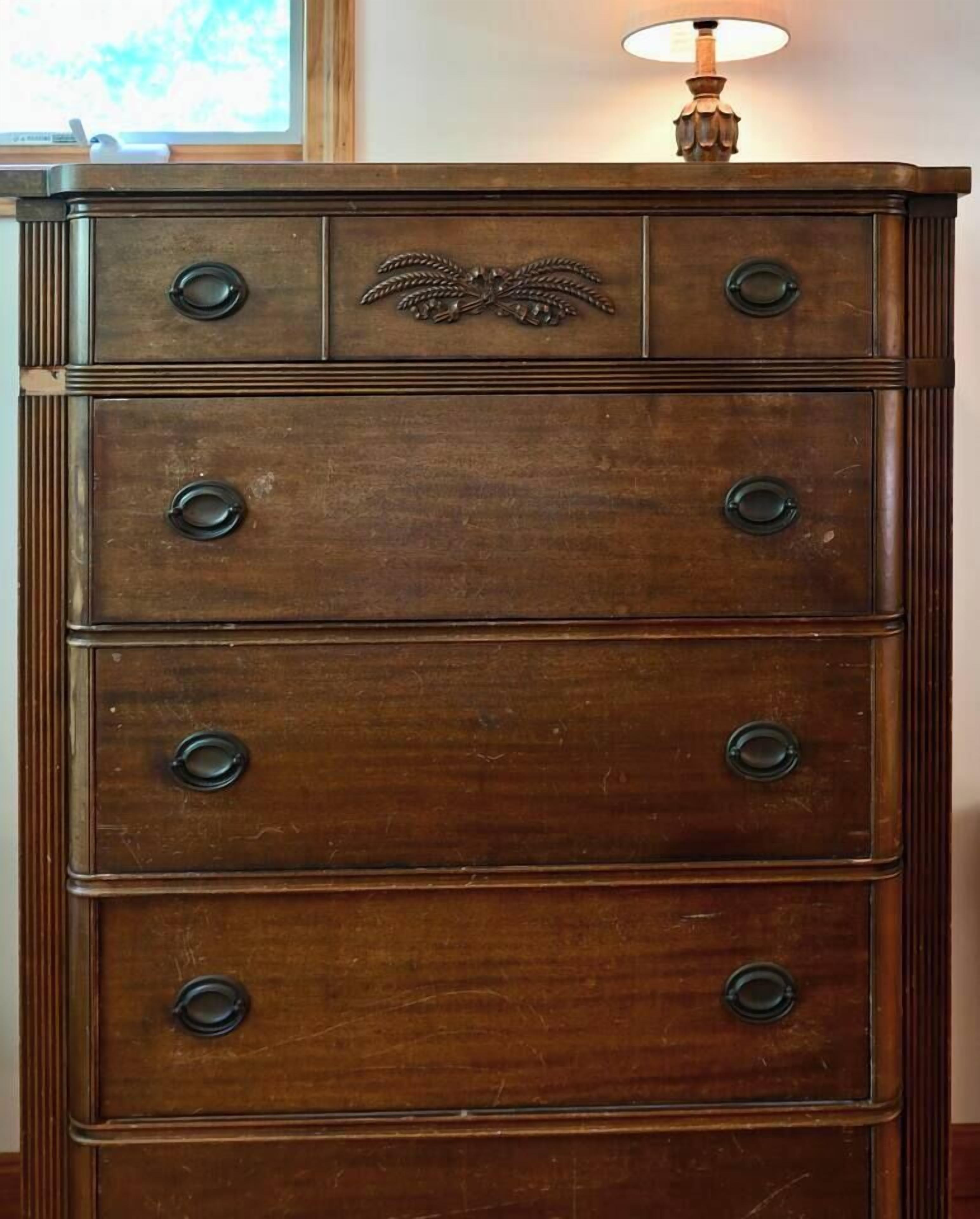 A wooden dresser with multiple drawers and knobs, a decorative emblem on the top, and a lamp on top.
