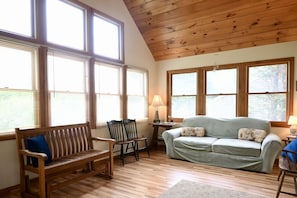 A cozy room with a wooden ceiling, large windows, a sofa, a bench, and a table with a lamp. - Beach n’ Bunks | Walk to Beach | Classic Cape (Sandwich)