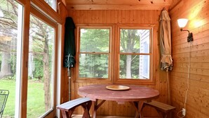 L’intérieur d’un chalet en bois, avec une table ronde, des bancs et de grandes fenêtres donnant sur une forêt.