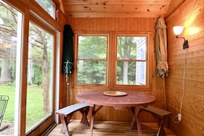 Interior de una cabaña de madera con una mesa redonda, bancos y grandes ventanales con vistas a un bosque.