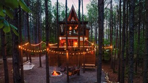 Una cabaña con terraza, fogata y luces colgantes en un entorno forestal.