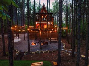 Eine Holzhütte mit Terrasse, Feuerstelle und Lichterketten in Waldumgebung.