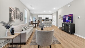 Een moderne woonkamer met een bank, fauteuil en een televisie. Er is een zithoek met een tafel en stoelen, en een keuken op de achtergrond.