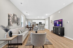 Una sala de estar moderna con sofá, sillón y televisor. Hay un comedor con mesa y sillas, y una cocina al fondo.