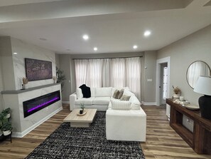 Ein modernes Wohnzimmer mit einem weißen Sofa, einem Kamin, einem Couchtisch und einem großen Fenster mit Vorhängen.