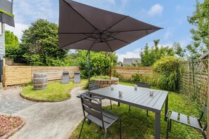 Eine Terrasse im Freien mit Tisch, Stühlen, einem großen Sonnenschirm und einem Steinfass.