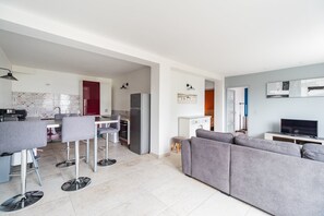 Ein zeitgemäßer Wohnbereich mit grauem Sofa, einer Essecke mit Barhocker, einer Küche mit roten Schränken und einem Fernseher auf einem Schrank.