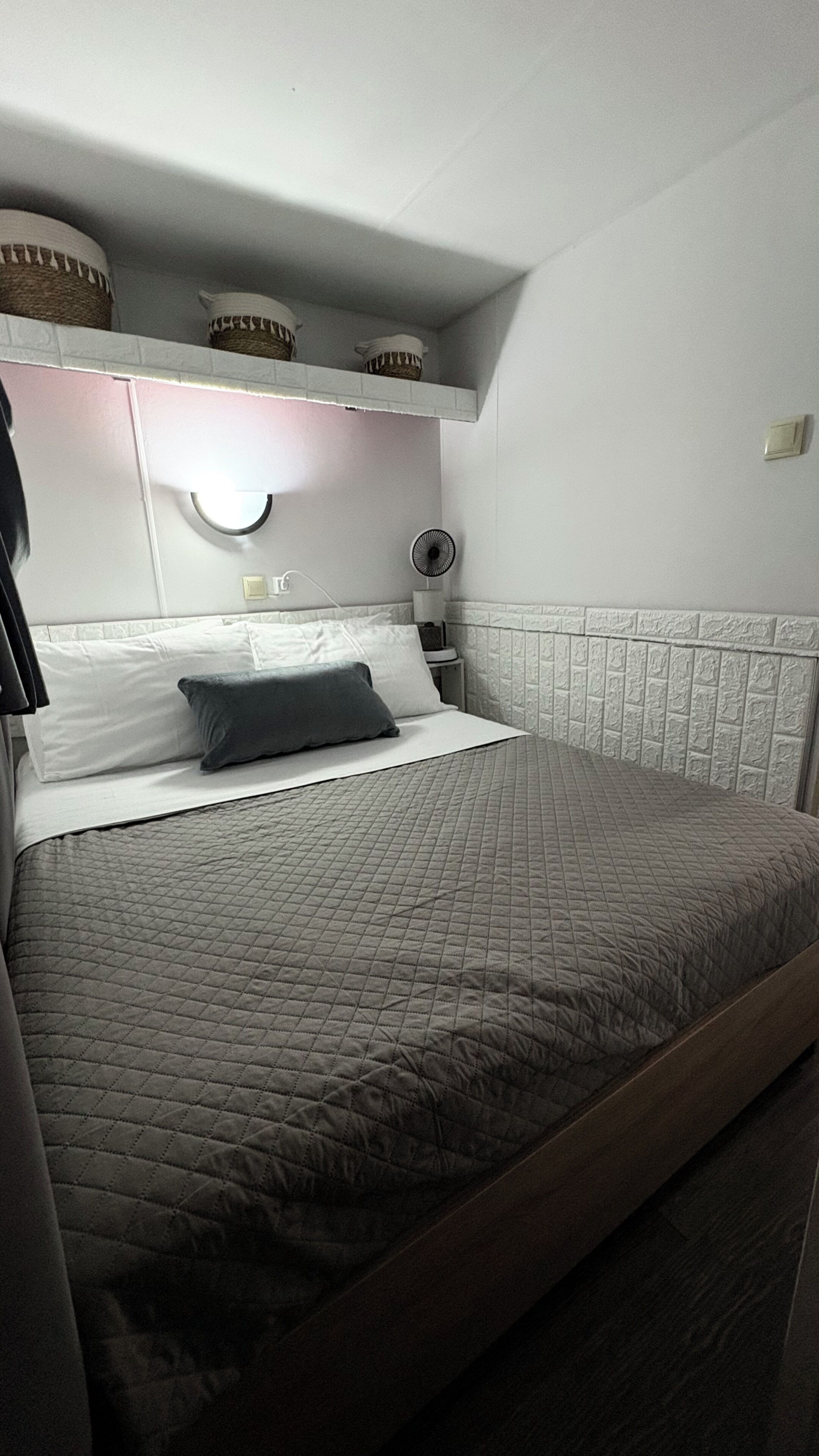 Una cama con colchón acolchado, una mesita de noche con lámpara y un ventilador de pared.