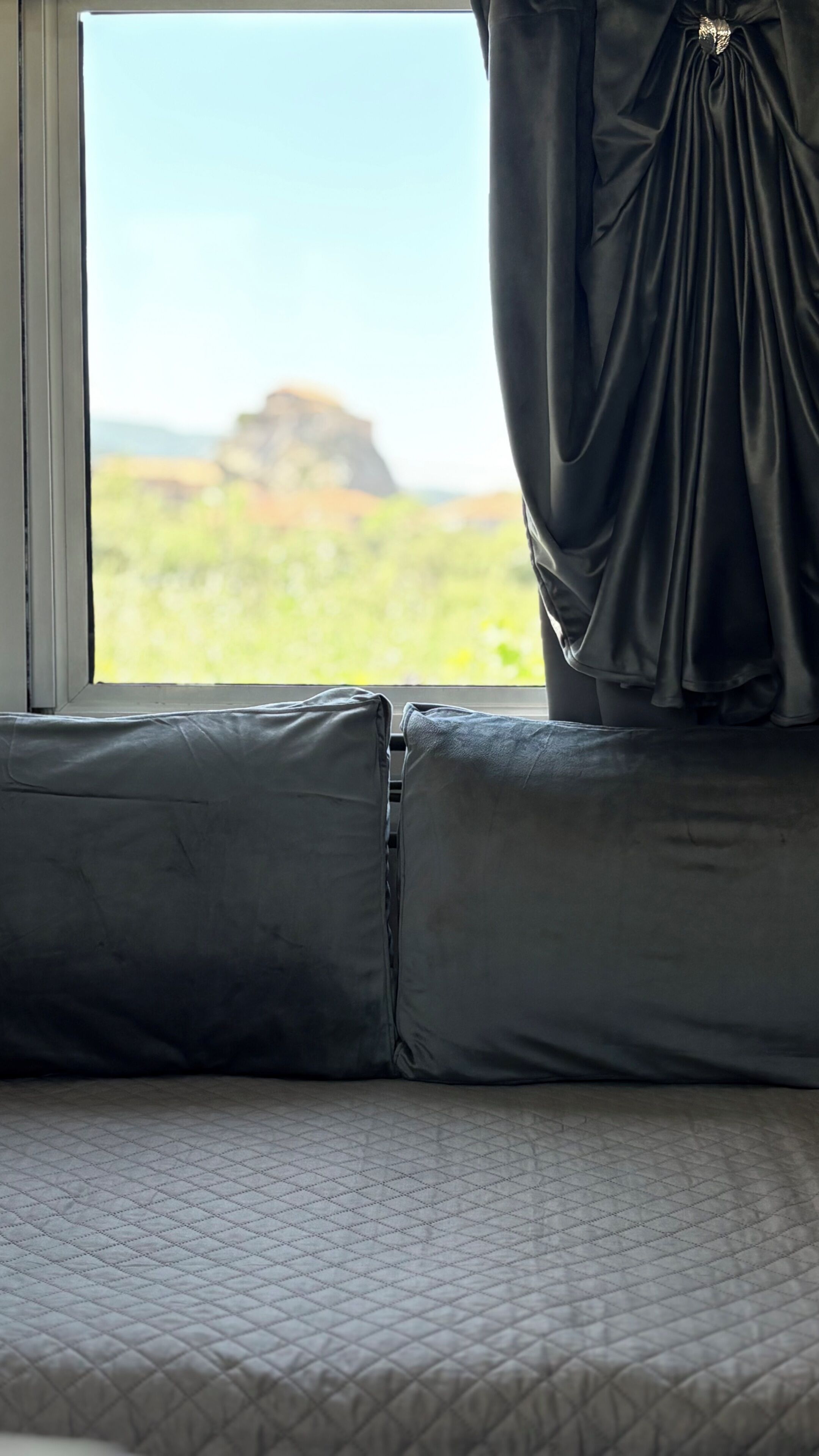Una cama con una colcha acolchada y almohadas, una ventana con vistas a un paisaje, y una cortina entreabierta.