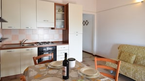 A compact kitchen with a dining table, chairs, a wine bottle, and a sofa in a small living area. - RES Magnolia 9 - LE Dimore Fiorite DI Comino (Siniscola)