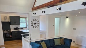 A modern kitchen with dark cabinets, a microwave, and an oven. There is a blue sofa with cushions, a wooden coffee table, and a dining table with chairs. - Boutique Dairy Conversion Near Ilford Station (Ilford)
