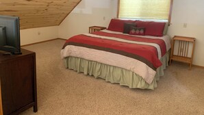 Une chambre avec un plafond en pente, un lit recouvert d’une couverture rouge et beige, un téléviseur posé sur un meuble en bois, et une fenêtre avec des stores.