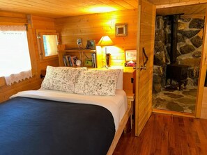A cozy log cabin bedroom with a bed, a nightstand, a lamp, a bookshelf, and a stone fireplace.