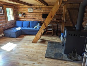 Interior de una cabaña de troncos con un sofá, una silla de madera, una chimenea y una escalera.
