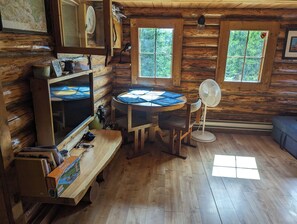 Interior de una cabaña de madera con una mesa redonda, un banco, un ventilador y una ventana con vistas a árboles.
