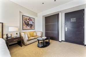 Ein Hotelzimmer mit einem Bett, einem Sofa, einem Couchtisch, einem Schreibtisch mit Lampe, einem Bild an der Wand und einer dunklen Tür.