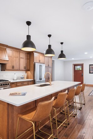 A modern kitchen with wooden cabinets, a stainless steel refrigerator, a large island with bar stools, and pendant lighting.