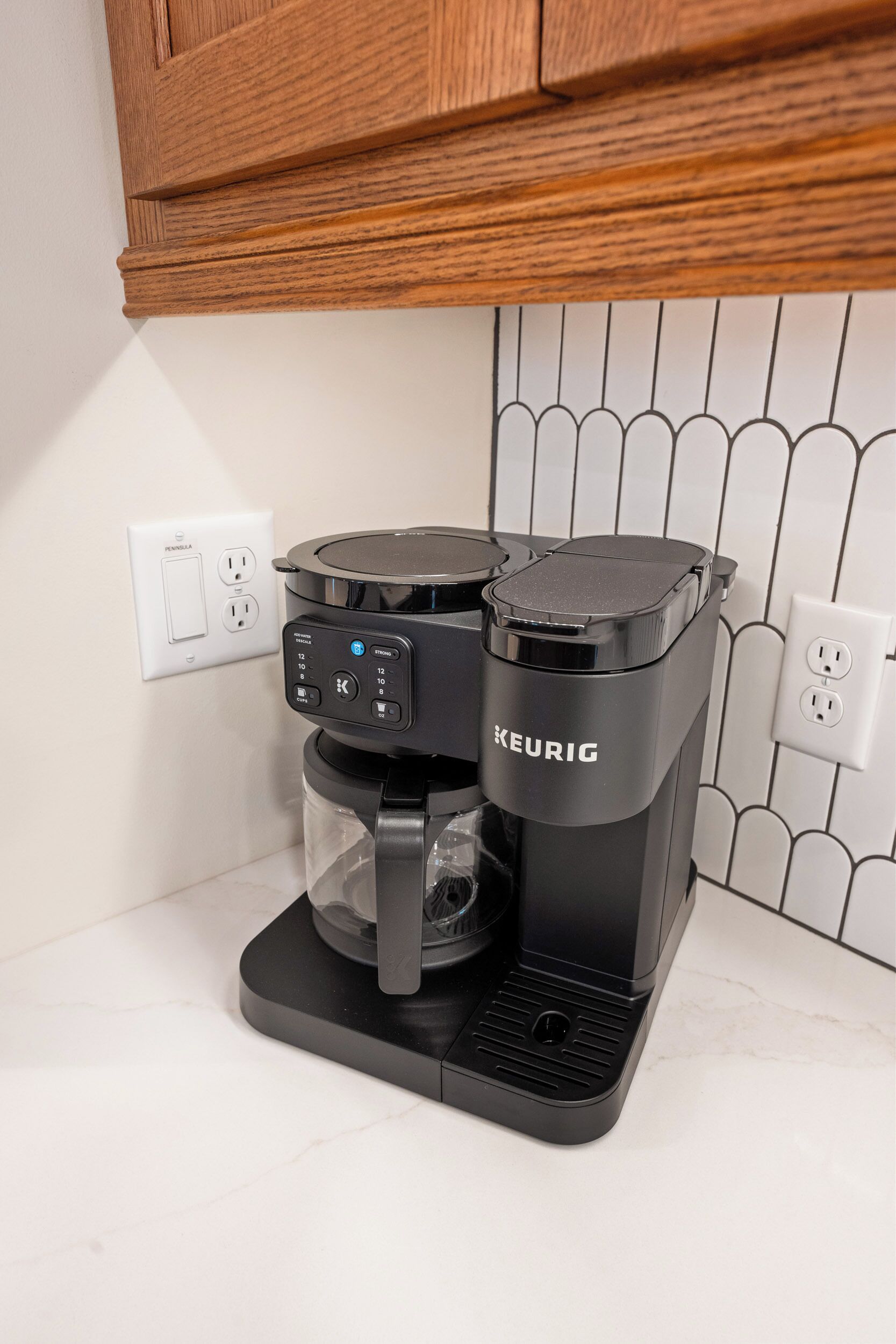 Una cafetera Keurig sobre una encimera con salpicadero blanco de azulejos y armarios de madera arriba.
