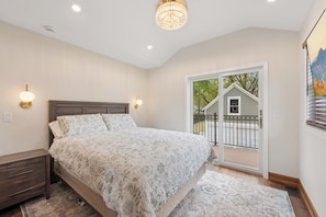 A bedroom with a large bed, a nightstand, a chandelier, and a view of a house through a sliding glass door.