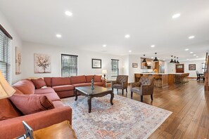 A spacious living area with a sectional sofa, a coffee table, and a patterned rug. There is a kitchen area with wooden cabinets and a bar.