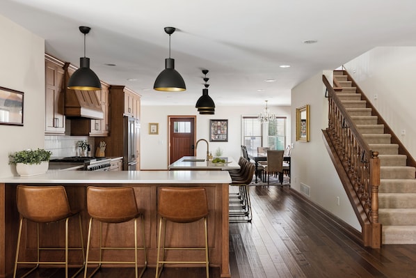 A modern kitchen with a central island, bar stools, pendant lighting, and a staircase leading to another level.