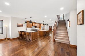 A spacious kitchen with wooden cabinets, a central island with bar stools, a staircase leading to an upper level, and recessed ceiling lighting.