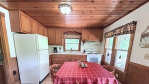 A kitchen with wooden cabinets, a white refrigerator, a table with a red checkered tablecloth, and a window with a view of greenery. - Cozy 1-bedroom cabin in perfect Wathena with AC (Wathena)
