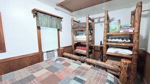 A room with a wooden bunk bed, a quilted bedspread, a window with blinds, and a shelf with folded items. - Cozy 1-bedroom cabin in perfect Wathena with AC (Wathena)