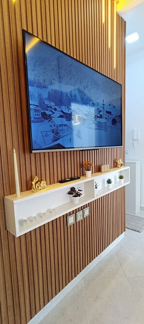 Ein modernes Wohnzimmer mit einem großen Flachbildfernseher an einer Holzwand, einem weißen Konsolentisch darunter und einem kleinen Regal mit Dekorationsobjekten.
