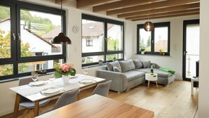 Eine moderne Küche mit Esstisch, Stühlen und einer Couch. Große Fenster bieten Ausblick nach draußen.