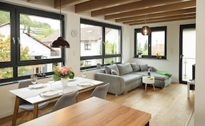 Eine moderne Küche mit Esstisch, Stühlen und einer Couch. Große Fenster bieten Ausblick nach draußen.
