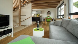 Ein modernes Wohnzimmer mit einem Flachbildfernseher, einer Couch, einem Couchtisch mit Flasche und Gläsern, einer Treppe und Blick auf die Küche.
