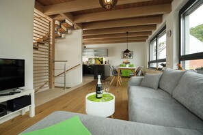Ein modernes Wohnzimmer mit einem Flachbildfernseher, einer Couch, einem Couchtisch mit Flasche und Gläsern, einer Treppe und Blick auf die Küche.