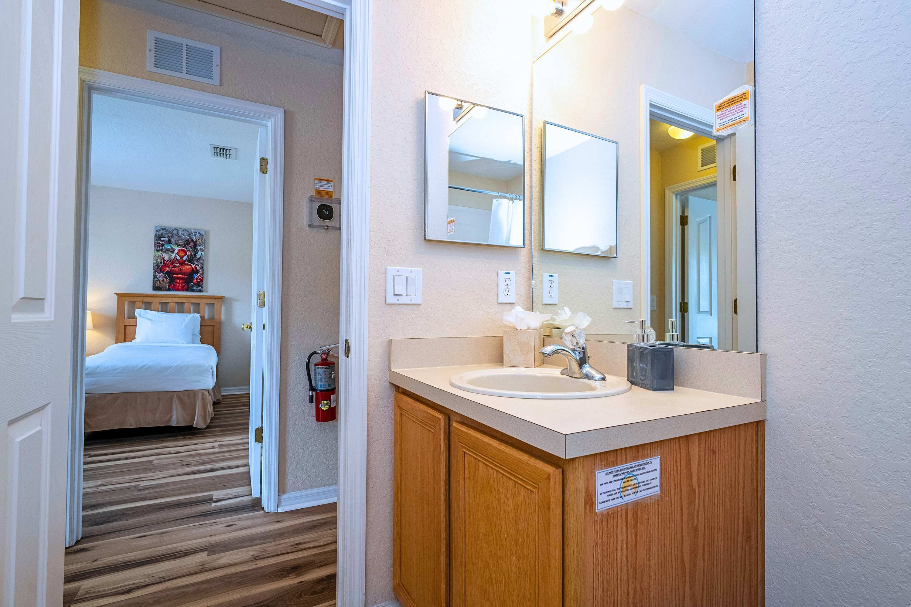 A hotel room with a wooden floor, a bed with white bedding, a bathroom with a sink, mirrors, and a fire extinguisher.