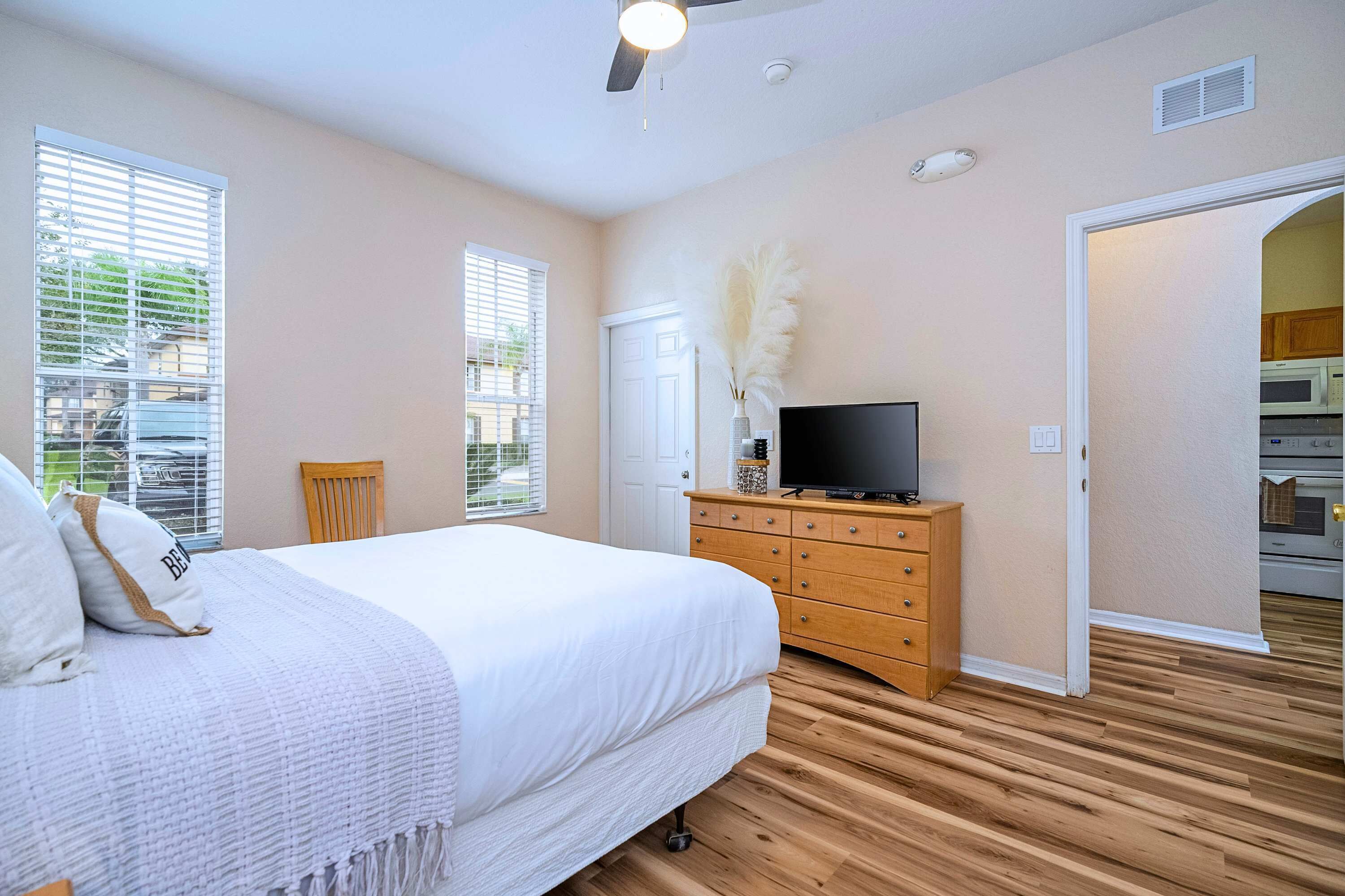A bedroom with a bed, wooden dresser, television, and a door leading to a kitchen.