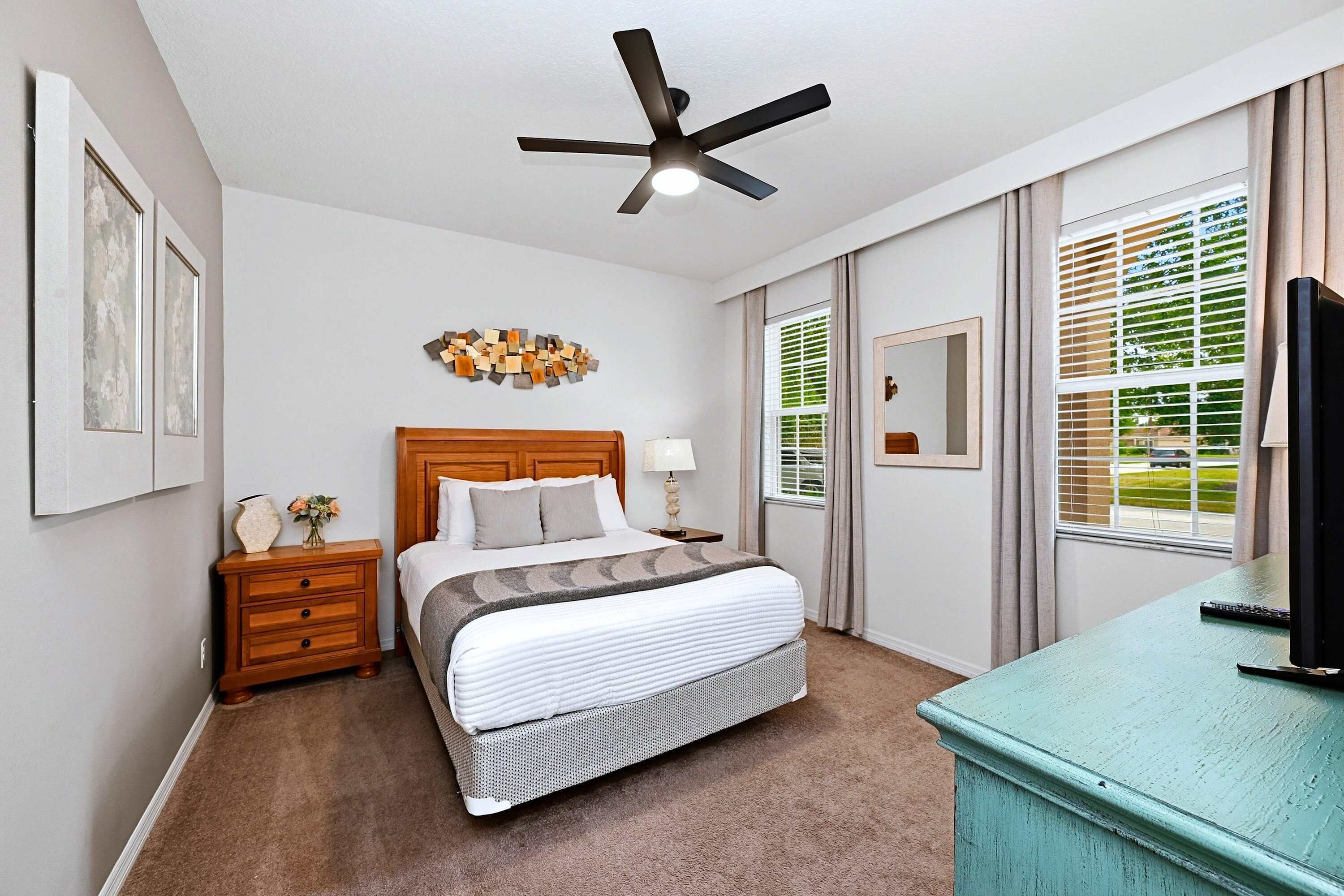 A bedroom with a bed, nightstand, lamp, ceiling fan, and a desk with a television.