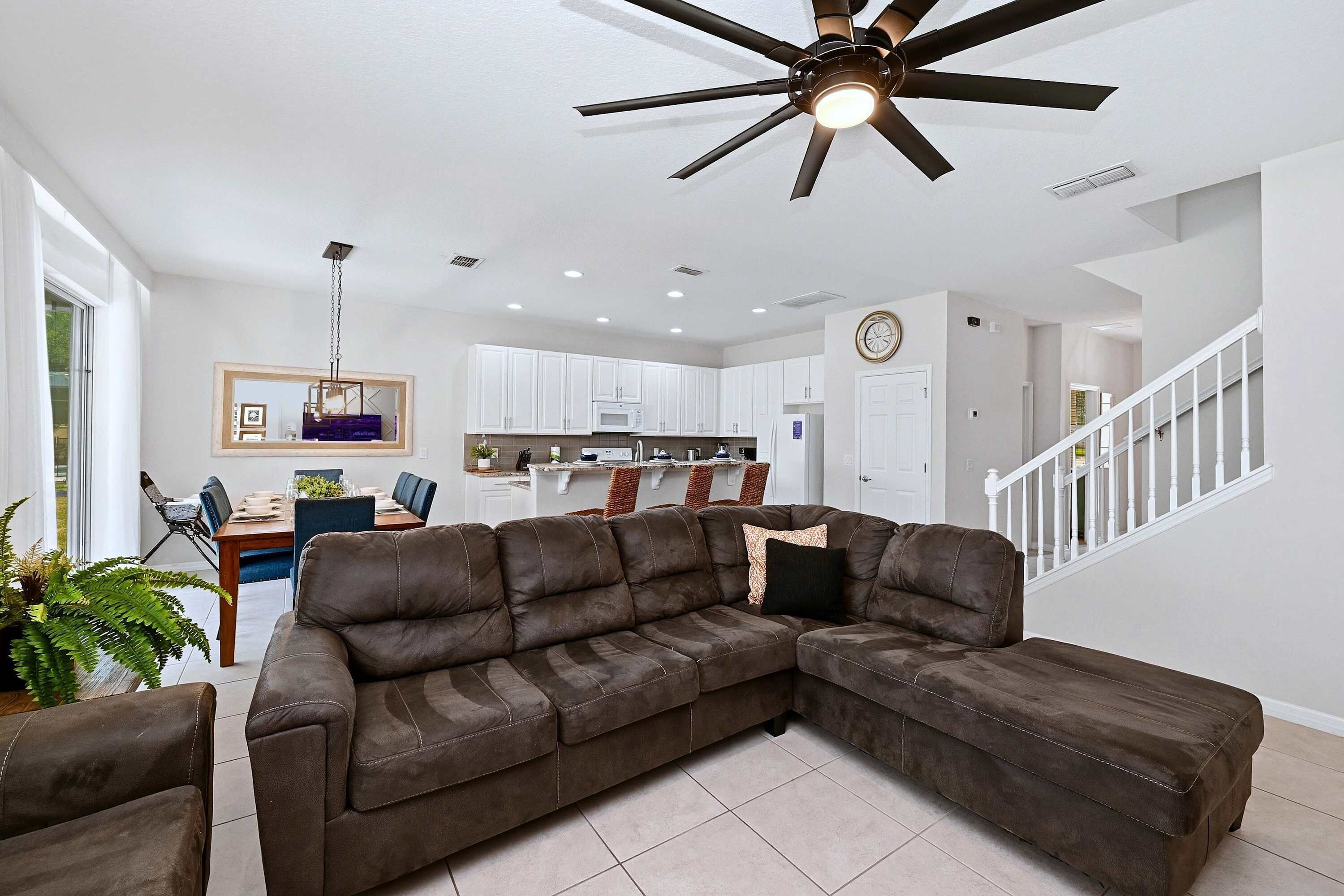 A living room with a sectional sofa, a ceiling fan, and a view to the kitchen.