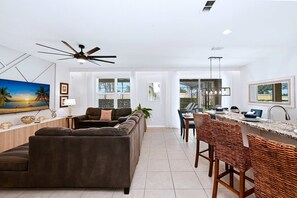 Ein modernes Wohnzimmer mit einer Couch, einem Essbereich mit Korbstühlen, einer Küche mit Bar und einem Deckenventilator.