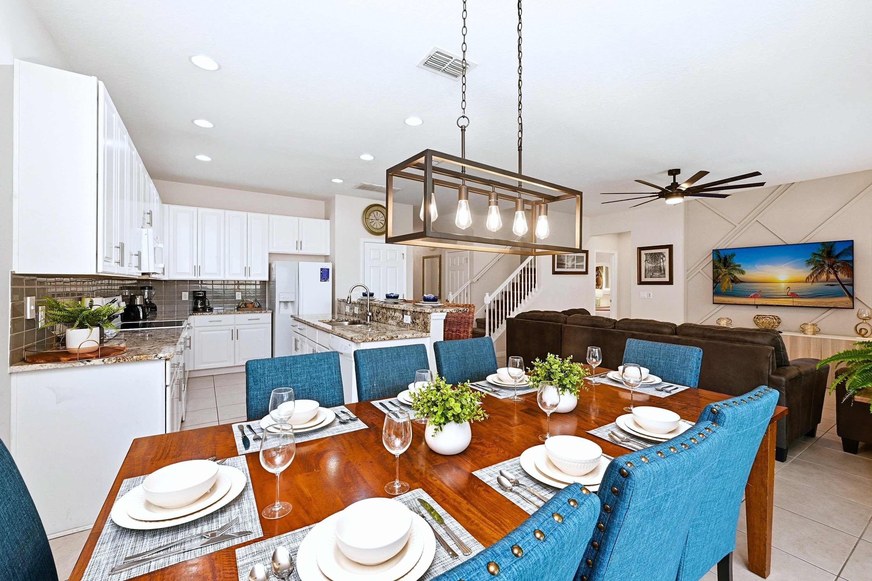 A modern kitchen with white cabinets, a dining area with a wooden table set for six, and a living room with a brown sofa and a ceiling fan.
