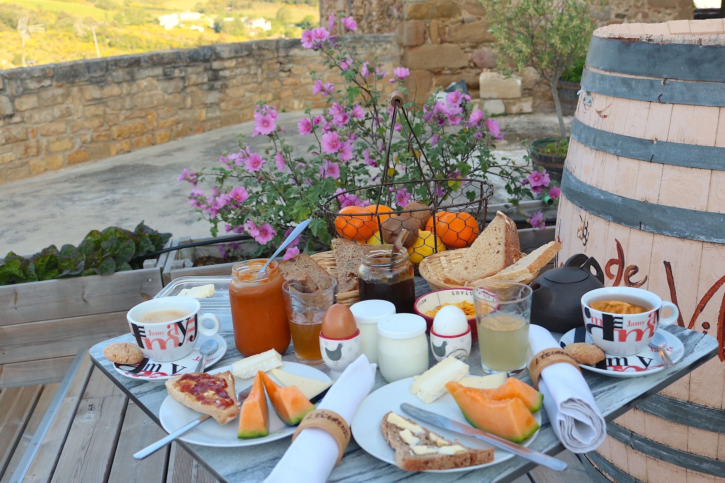 Une table dressée à l’extérieur, avec de la nourriture et des boissons, notamment des sandwichs, des fruits et diverses boissons, et offrant une vue sur un mur de pierre et des fleurs.