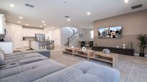 Ein modernes Wohnzimmer mit grauem Sofa, einem an der Wand montierten Fernseher und einer Küche im Hintergrund.