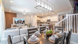 A modern living room with a flat-screen TV, a dining area with a table set for six, and a kitchen in the background. - Luxury 5BR House Private Pool & BBQ Sleeps 18 (Davenport)