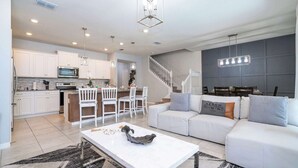 A modern kitchen with white cabinets, a dining area with white chairs, a white sofa, and a staircase. - Luxury 5BR House Private Pool & BBQ Sleeps 18 (Davenport)