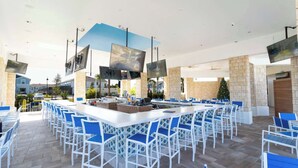 Uma área de bar espaçosa ao ar livre, com diversas opções de assentos e televisores de tela plana fixados nas paredes.