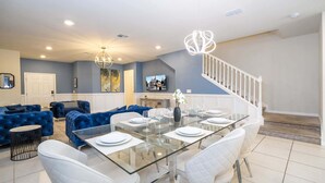 Uma sala de jantar moderna com uma mesa de vidro posta para seis pessoas, um sofá azul, um candelabro e uma escada.