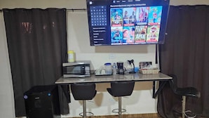 A small kitchenette with a microwave, coffee maker, and a toaster. There are two black bar stools and a TV mounted on the wall displaying movie recommendations. - Tocumen Comfort Stay (24 de Diciembre)