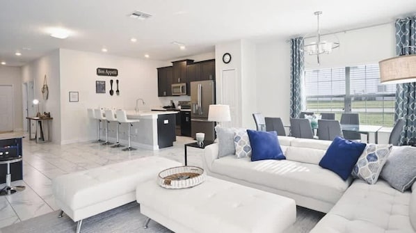 Una sala de estar moderna con un sofá blanco, un comedor con sillas, una cocina con armarios oscuros y un ventanal grande.