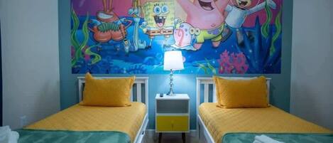 A room with two beds, a mural of cartoon characters on the wall, and a nightstand with a lamp.