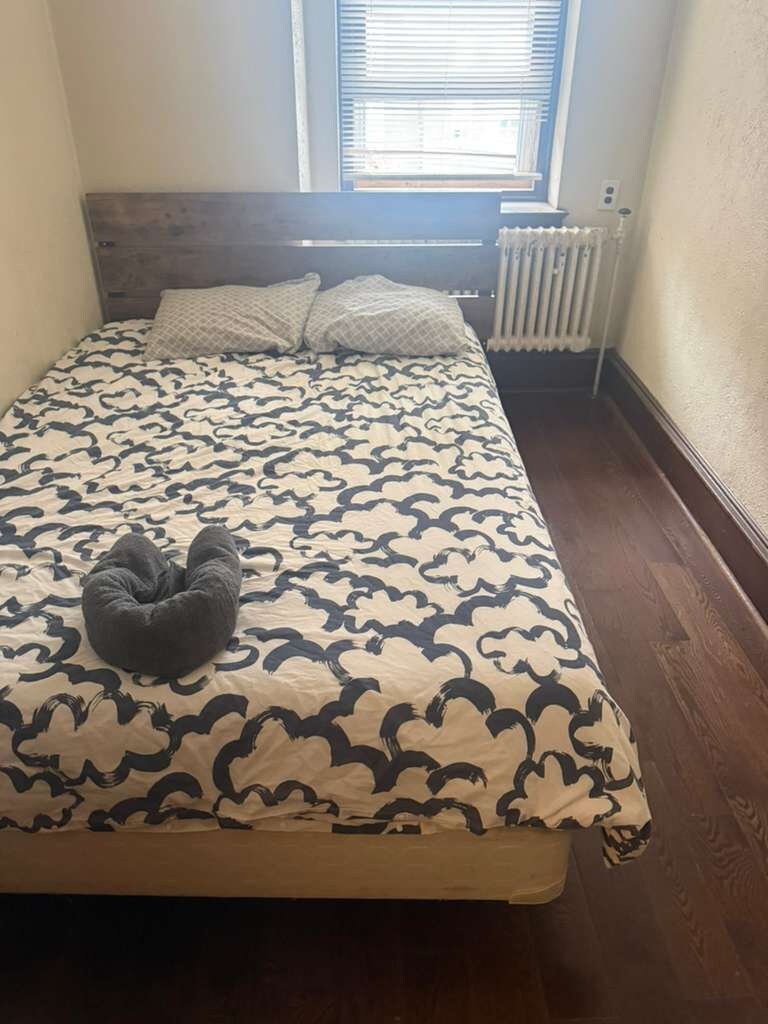 Un dormitorio con cama, ventana con persianas, radiador y almohada oscura.