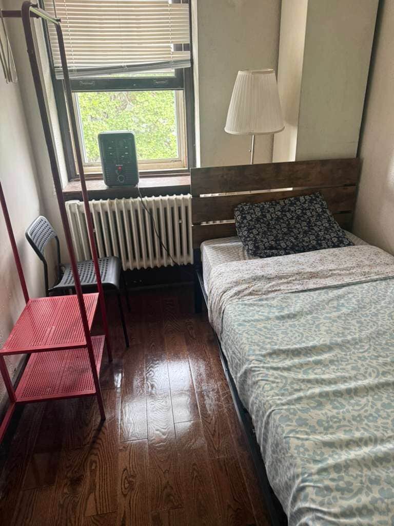 A room with a bed, a chair, a radiator, and a window with blinds.